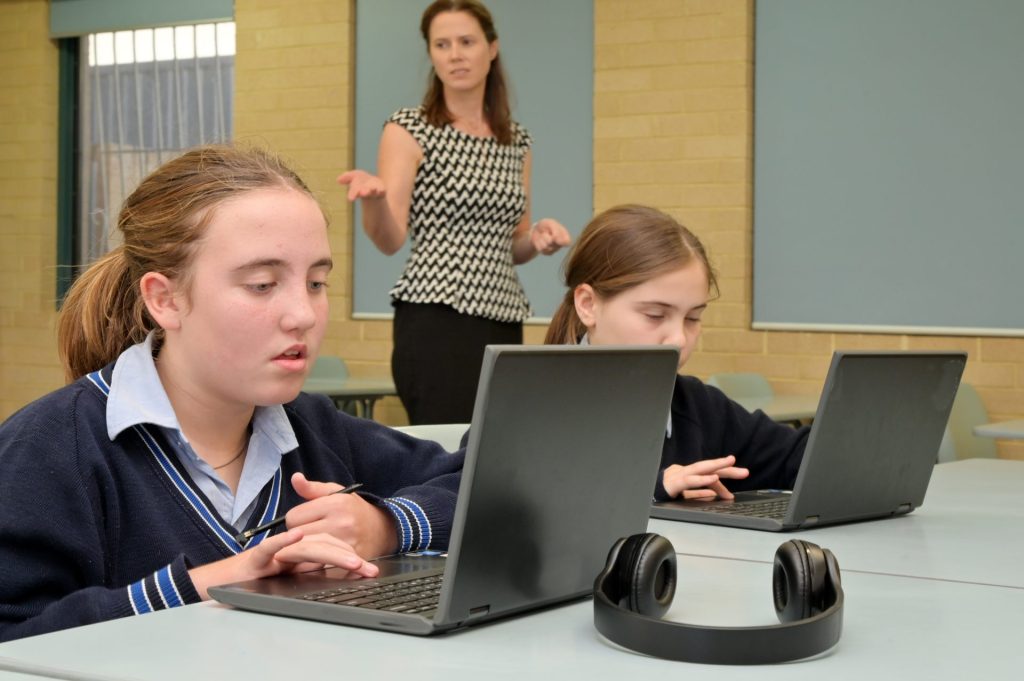 2 teenagers in school uniform using laptops. There is a teacher in the background giving instructions.